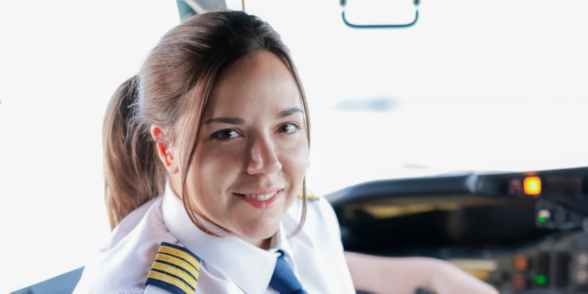 Une pilote souriante est assise dans le cockpit, portant une chemise blanche et des épaulettes dorées. Des instruments de vol sont visibles en arrière-plan.